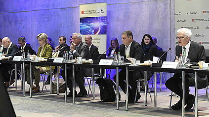 Die baden-württembergischen Minister:innen Winfried Hermann, Dr. Nicole Hoffmeister-Kraut, Thomas Strobl, Winfried Kretschmann und e-mobil BW Geschäftsführer Franz Loogen sitzen an einem langen Konferenztisch mit Mikrofonen und Namensschildern. Im Hintergrund sind Zuschauende zu sehen. 